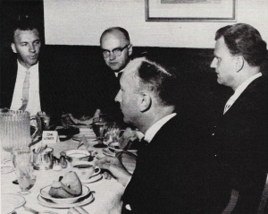 Black and white photo of four men sitting around a table with food on it.