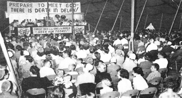 A large crowd of people sit under a large canvas tent watching a preacher behind a podium. Large signs are posted behind him, proclaiming Christian messages.